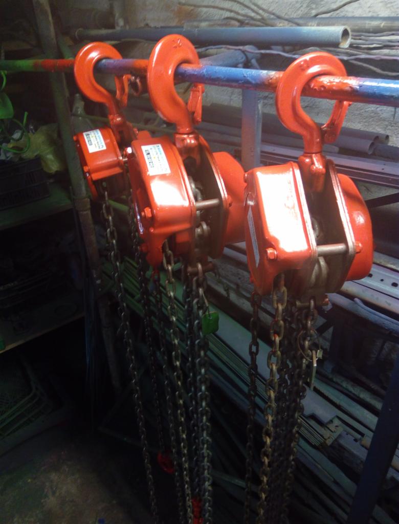 Trzy czerwone wciągniki łańcuchowe zawieszone na niebieskiej rurze, z łańcuchami zwisającymi w dół, w tle widoczne półki z metalowymi elementami.