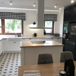 Jasna kuchnia z białymi szafkami, drewnianym blatem, wyspą kuchenną, czarno-białymi roletami w kratę i wzorzystą podłogą w stylu retro, widziana z perspektywy jadalni.