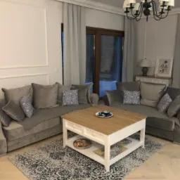 Salon w odcieniach szarości z dwoma welurowymi sofami, stolikiem kawowym z drewnianym blatem i białymi nogami, dywanem w stylu vintage i dekoracyjnymi poduszkami w niebieski wzór.