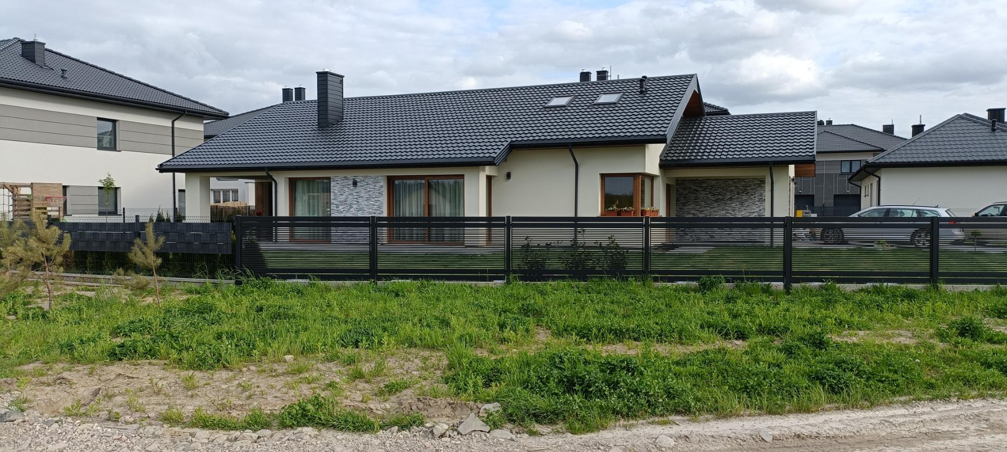 Nowoczesny dom jednorodzinny z ciemną dachówką i elewacją w jasnym odcieniu, widoczny zza czarnego, poziomego ogrodzenia panelowego.