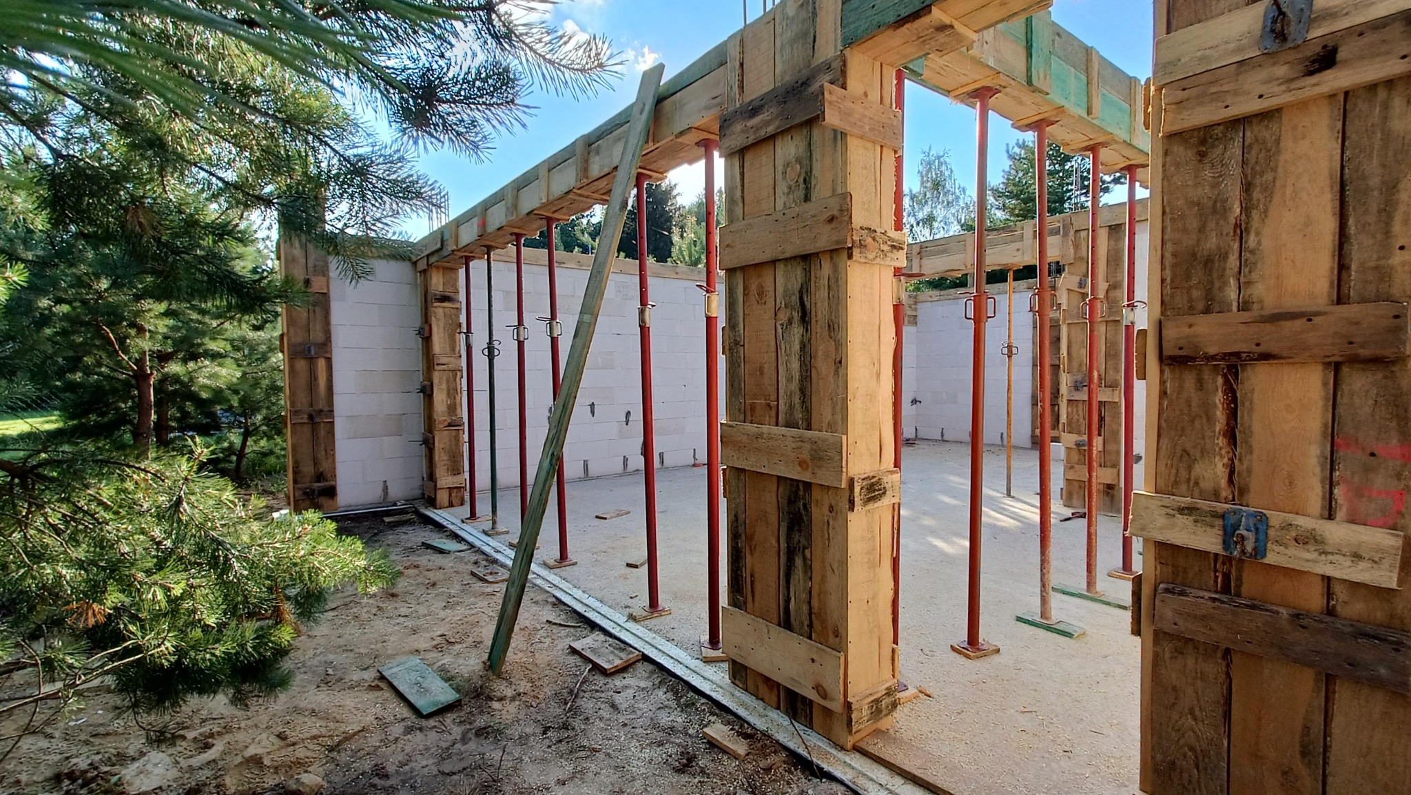 Wznoszenie ścian z bloczków betonowych z drewnianym szalunkiem i stalowymi podporami, widok z zewnątrz na tle zieleni.