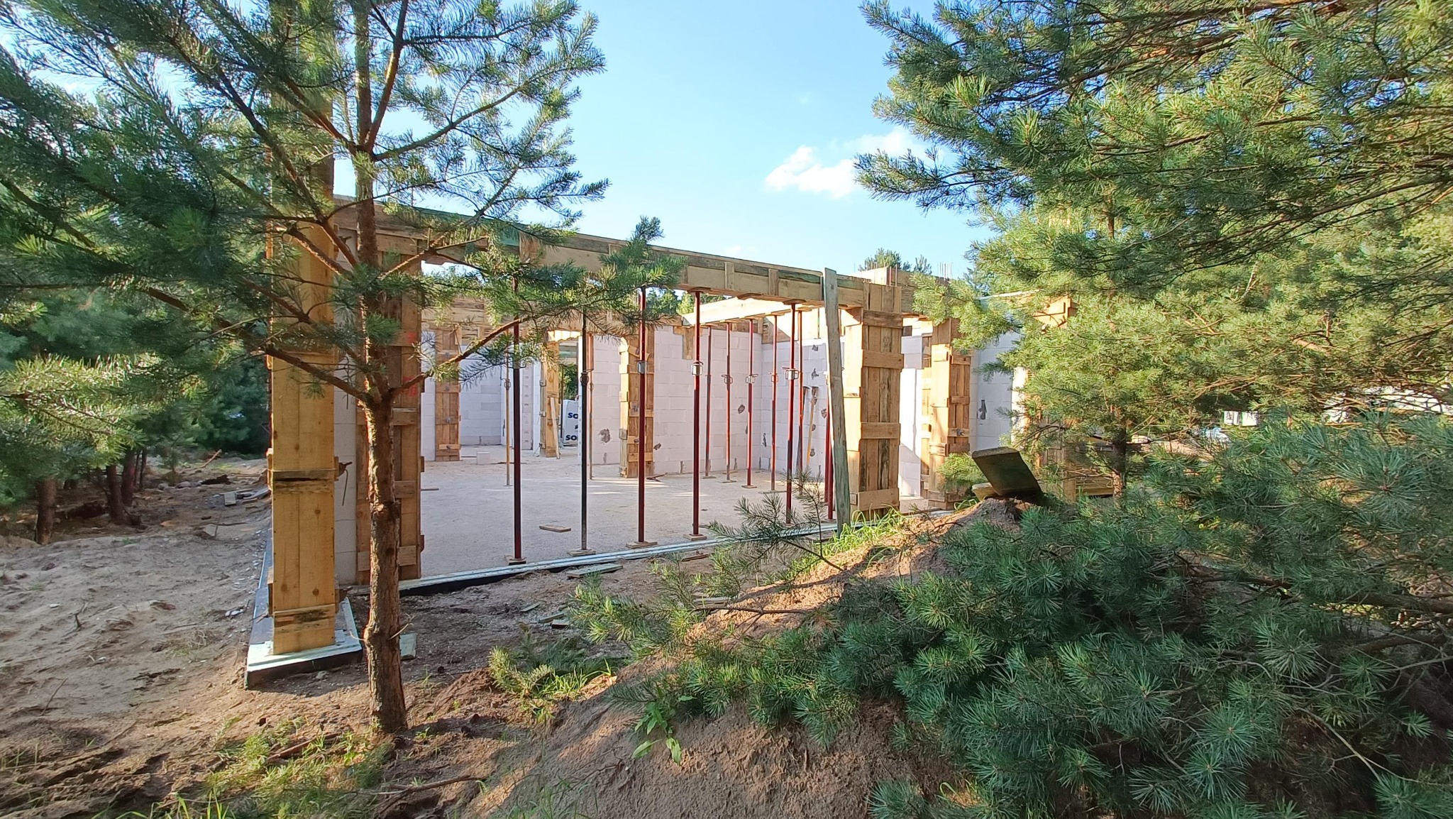 Surowa konstrukcja domu w lesie, widoczne białe ściany z betonu komórkowego, drewniane szalunki i podpory, ujęcie zza drzew iglastych.