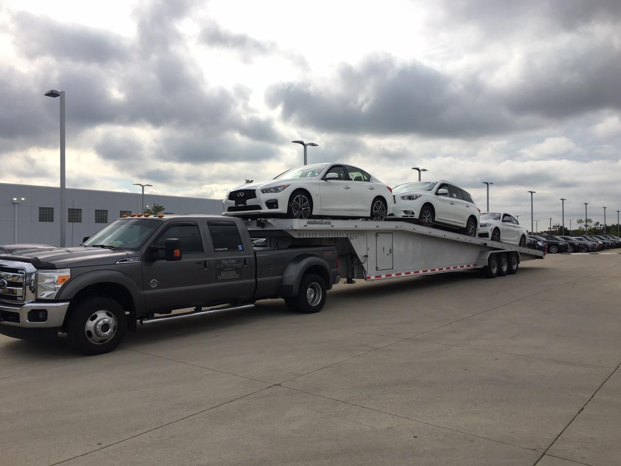 Szara laweta z trzema białymi samochodami osobowymi marki Infiniti, zaparkowana na betonowym placu, na tle budynku i pochmurnego nieba.