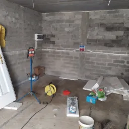Wnętrze surowego pomieszczenia z widoczną instalacją elektryczną: białe kable prowadzone po ścianie z bloczków, skrzynka elektryczna, gniazdka, lampa robocza na trójnogu, zwinięty przedłużacz,...