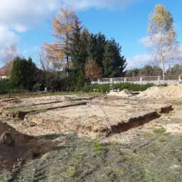 Wykopane rowy pod fundamenty domu jednorodzinnego, widoczne przygotowane zbrojenia i instalacje, w tle drzewa i domy.