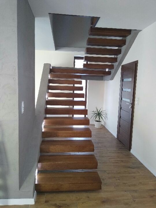 Nowoczesne, drewniane schody dywanowe z oświetleniem punktowym na ścianie, prowadzące na piętro, widok z dołu na tle parkietu i drzwi wewnętrznych.