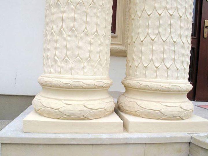 Dwie kremowe, zdobione kolumny z motywem łusek, ustawione na kwadratowych podstawach, z widocznymi fragmentami drzwi i okna w tle.