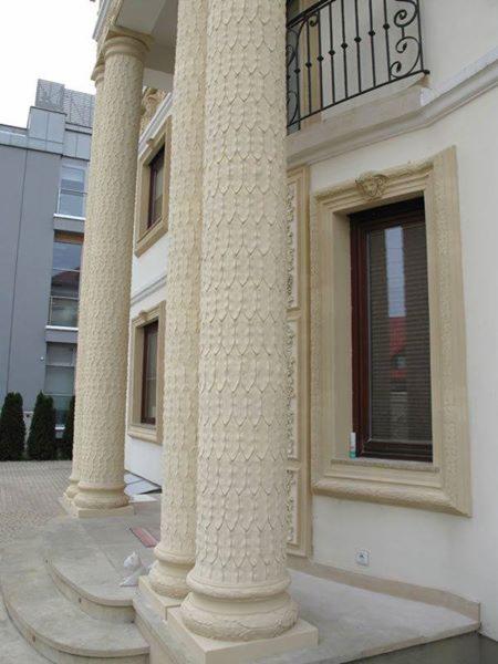 Elewacja budynku z kolumnami o dekoracyjnej fakturze łusek, ozdobne obramowanie okien z motywem twarzy, balkon z kutą balustradą.