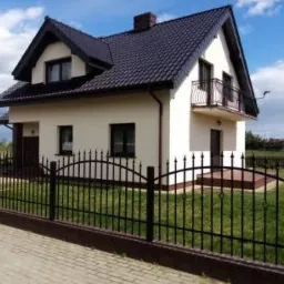 Dwukondygnacyjny dom jednorodzinny z balkonem, wykończony jasnym tynkiem, z ciemną dachówką i ozdobnym, metalowym ogrodzeniem frontowym na tle zielonego trawnika i błękitnego nieba.