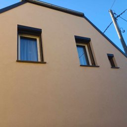 GREG-POL - Elewacja domu jednorodzinnego w odcieniach beżu z trzema oknami o czarnych ramach i roletach. Widok z dołu, na tle błękitnego nieba i słupa energetycznego.