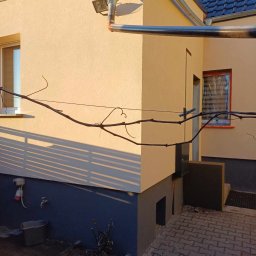 GREG-POL - Elewacja domu po tynkowaniu i malowaniu na beżowo-szaro. Widoczne rynny, okna z brązowymi ramami, fragment dachu z dachówką i kostka brukowa.