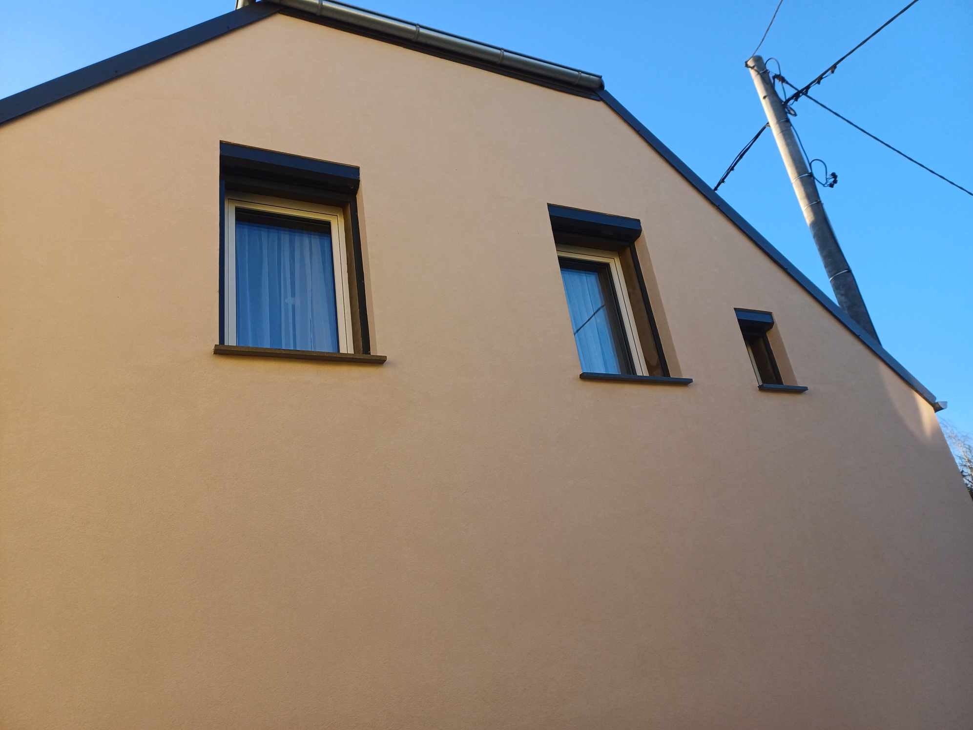 Elewacja domu jednorodzinnego w odcieniach beżu z trzema oknami o czarnych ramach i roletach. Widok z dołu, na tle błękitnego nieba i słupa energetycznego.
