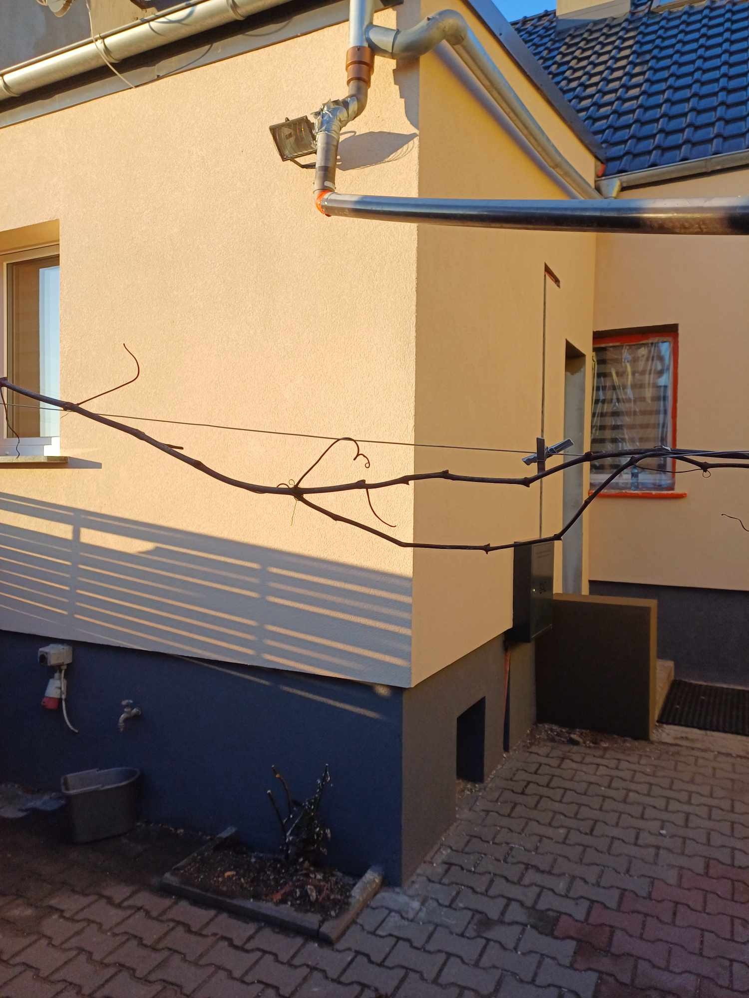 Elewacja domu po tynkowaniu i malowaniu na beżowo-szaro. Widoczne rynny, okna z brązowymi ramami, fragment dachu z dachówką i kostka brukowa.