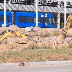 Dwa żółte koparki CAT pracują na placu budowy, w tle niebieski tramwaj i konstrukcja hali.