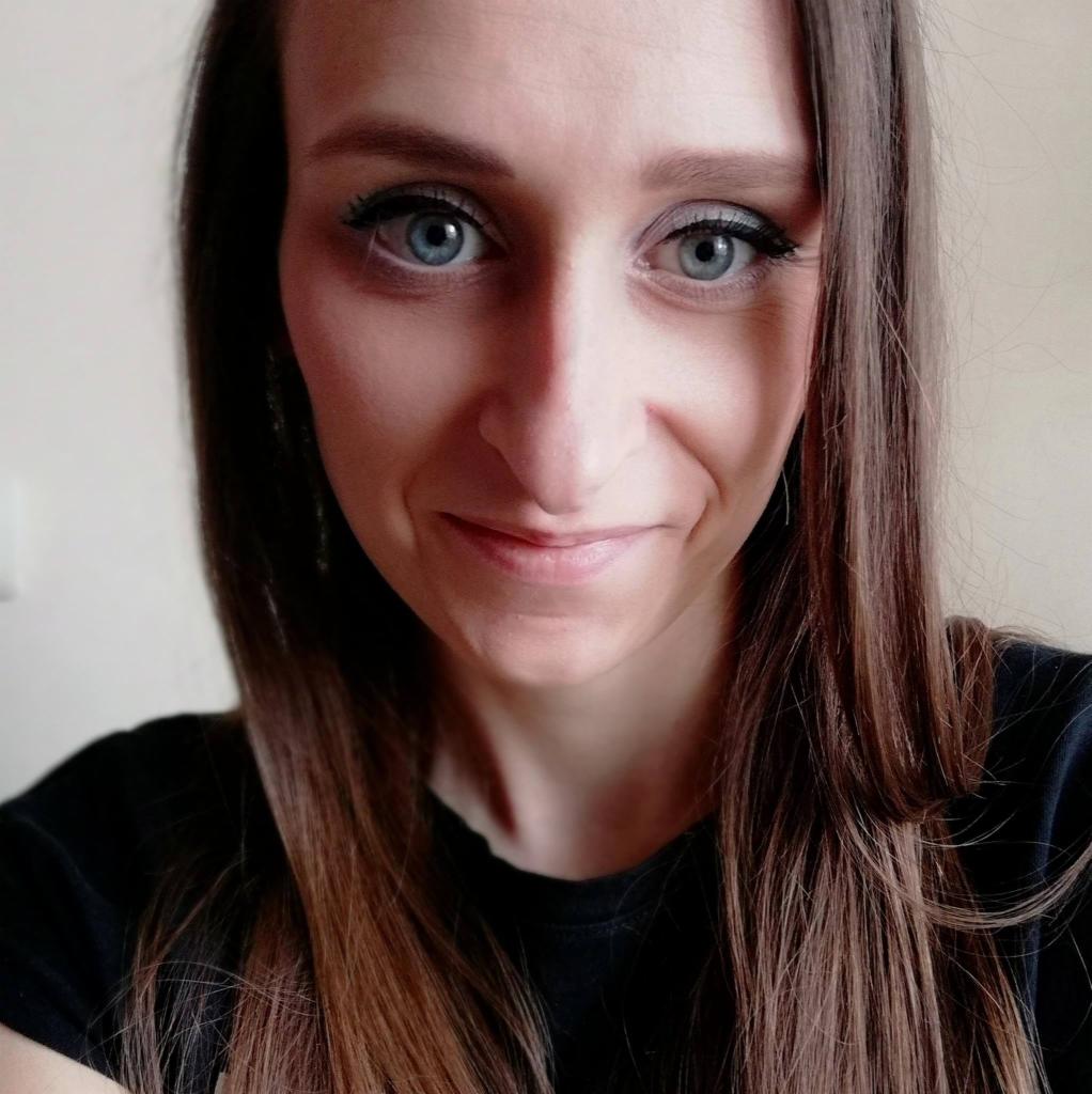 Portret uśmiechniętej kobiety z długimi brązowymi włosami i intensywnie niebieskimi oczami, ubrana w czarną koszulkę, na jasnym tle.