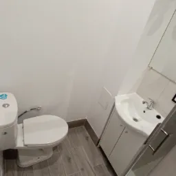 Nowoczesna, minimalistyczna łazienka z białą ceramiką, szarą podłogą imitującą drewno i elementami kabiny prysznicowej.