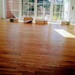 Nowo ułożona drewniana podłoga w jasnym pomieszczeniu z dużymi oknami, widoczne kartony po panelach i osoba pracująca na zewnątrz.