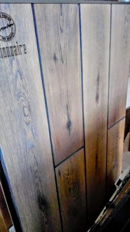 Ułożone panele podłogowe w odcieniach brązu i beżu, z widocznym usłojeniem drewna i ciemnymi fugami, na jednym z paneli widoczny napis 'Millionaire' z logo.