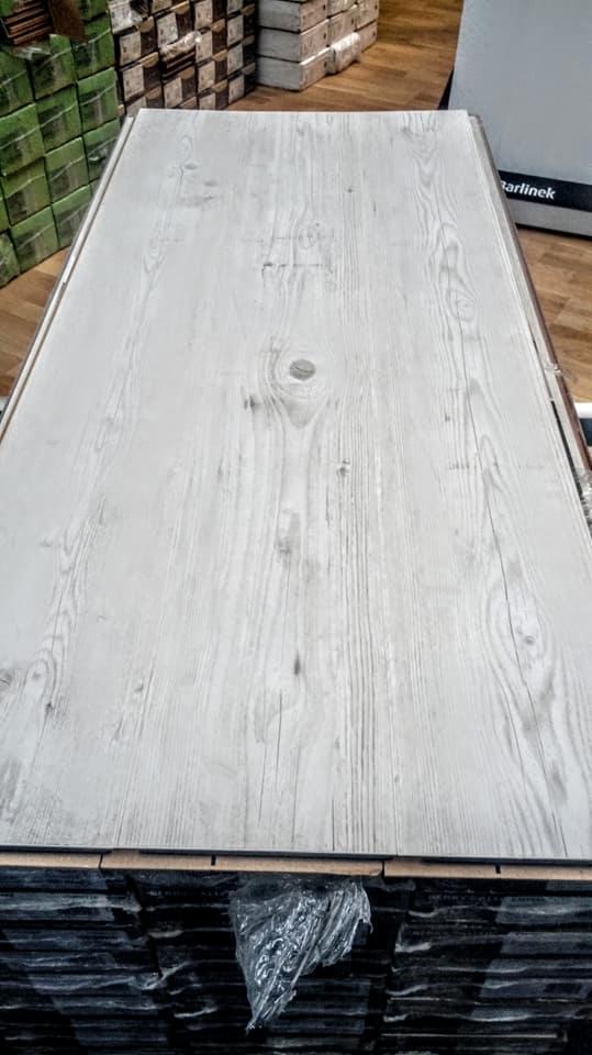 Stos paneli podłogowych w odcieniach szarości z widoczną strukturą drewna, opakowanych w folię, z logo 'Tarlinek' widocznym w tle.