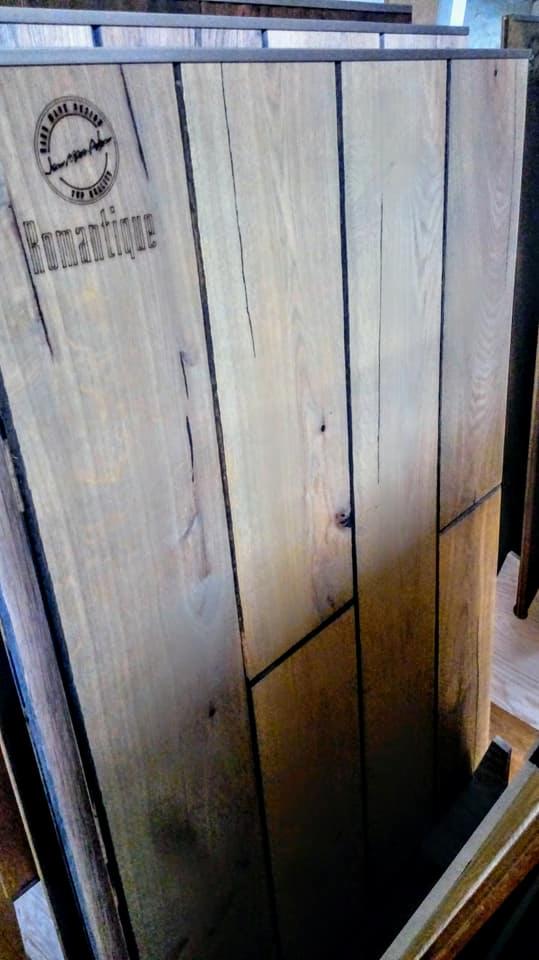 Próbka podłogi drewnianej z widocznym logo i nazwą 'Romantique' oraz naturalnymi pęknięciami i sękami w drewnie.