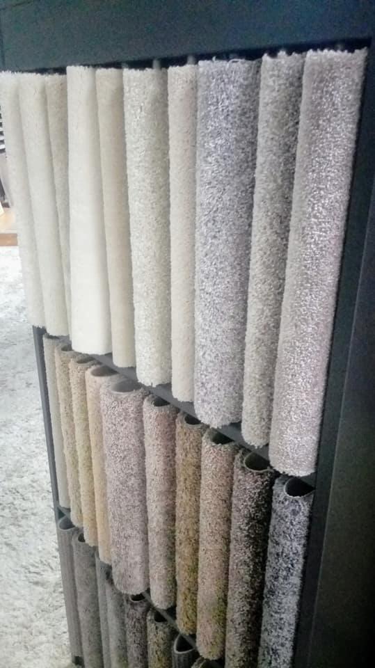 Ekspozycja próbek wykładzin dywanowych w różnych odcieniach bieli, beżu i szarości, ułożonych w rzędach na metalowym stojaku.