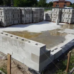 Budowa domu z keramzytu: widok na fundamenty i pierwsze warstwy ścian z bloczków keramzytowych, palety z materiałem budowlanym w tle, teren budowy po deszczu.