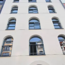 Fasada budynku z oknami o różnym kształcie i kolorze ram, w tym okna łukowe z szarymi ramami i okna prostokątne z czarnymi ramami, oraz fragment elewacji wykończony drewnem.