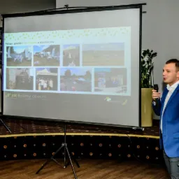 Prezentacja firmy na ekranie projekcyjnym: prelegent w niebieskiej marynarce z mikrofonem, zdjęcia z pikników i dni otwartych dla dostawców, logo firmy Aureus w prawym dolnym rogu ekranu.