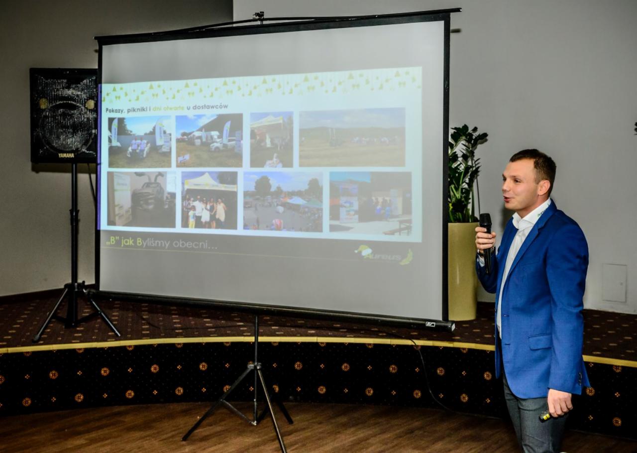 Prezentacja firmy na ekranie projekcyjnym: prelegent w niebieskiej marynarce z mikrofonem, zdjęcia z pikników i dni otwartych dla dostawców, logo firmy Aureus w prawym dolnym rogu ekranu.