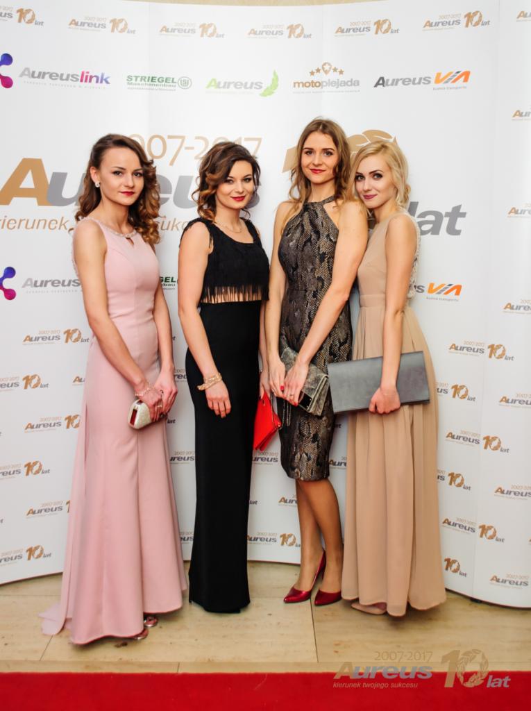 Cztery elegancko ubrane kobiety pozują na tle ściany z logo Aureus i napisami '10 lat', trzymając małe torebki.