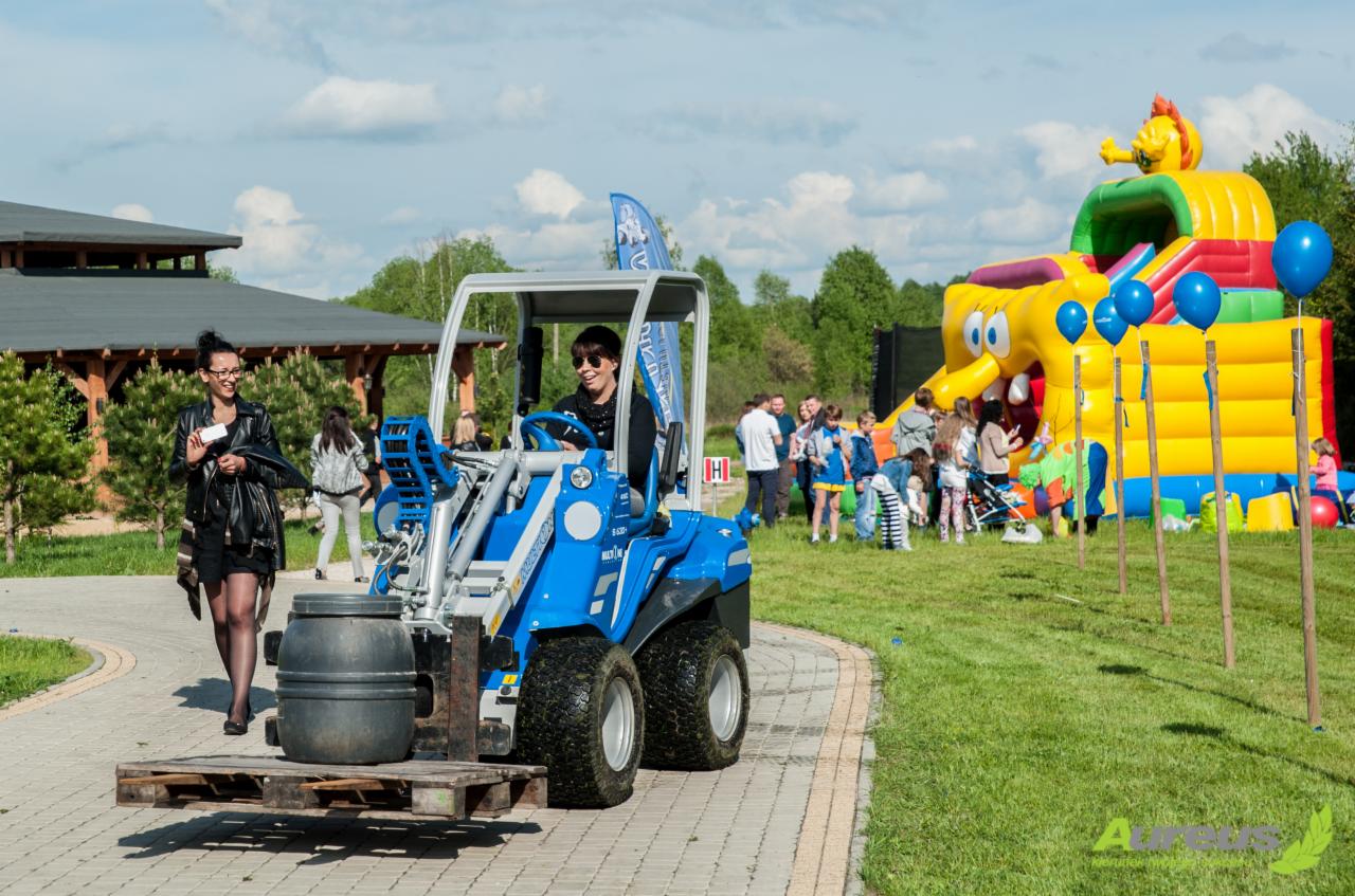 Kompaktowa, niebieska ładowarka teleskopowa przewozi beczkę na palecie po brukowanej ścieżce, w tle dmuchany zamek i grupa osób.