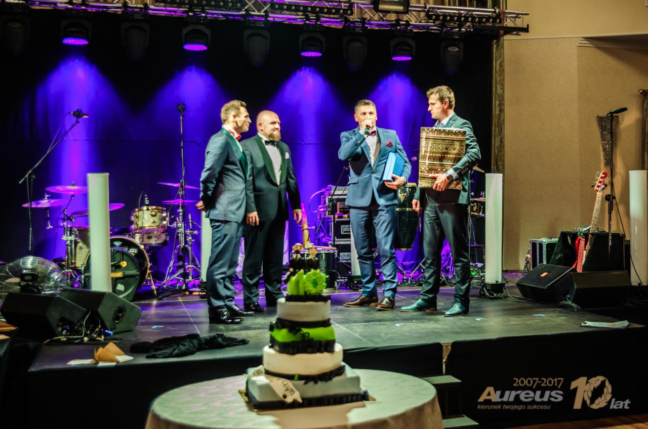 Scena z czterema mężczyznami w garniturach podczas uroczystości, tort na pierwszym planie, instrumenty muzyczne i oświetlenie sceniczne w tle.