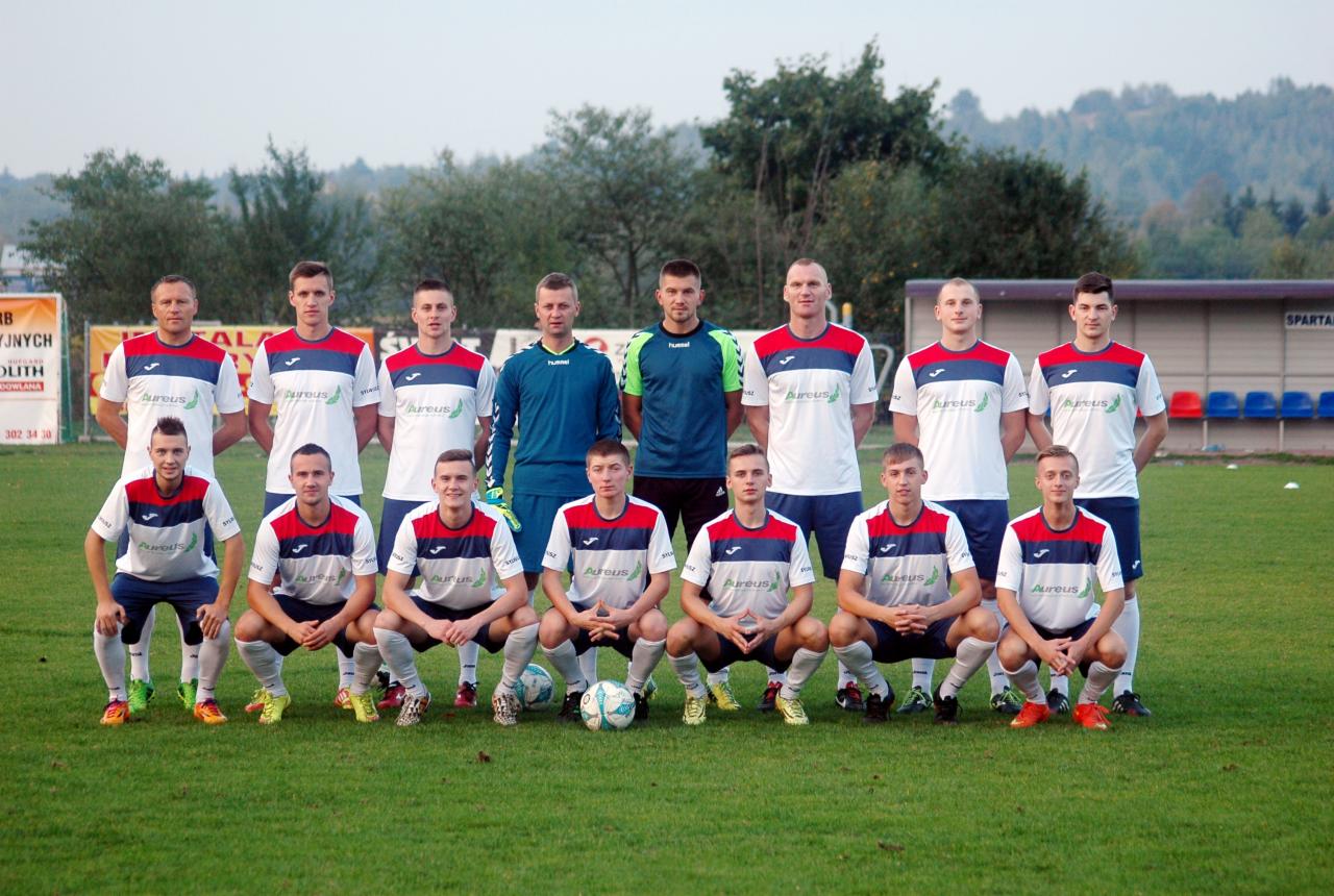 Drużyna piłkarska w strojach z logo sponsora 'Aureus' pozuje do zdjęcia na zielonym boisku.
