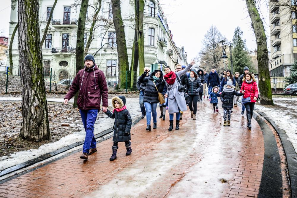 Grupa ludzi, w tym mężczyzna trzymający za rękę małą dziewczynkę, idzie po mokrym chodniku wzdłuż ulicy z budynkami i drzewami w tle.