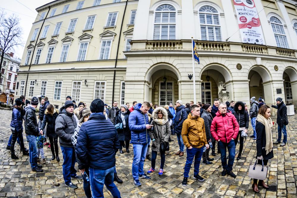 Tłum ludzi na brukowanym placu przed zabytkowym budynkiem z kolumnami i flagą, w tle drzewo bez liści i baner reklamowy.