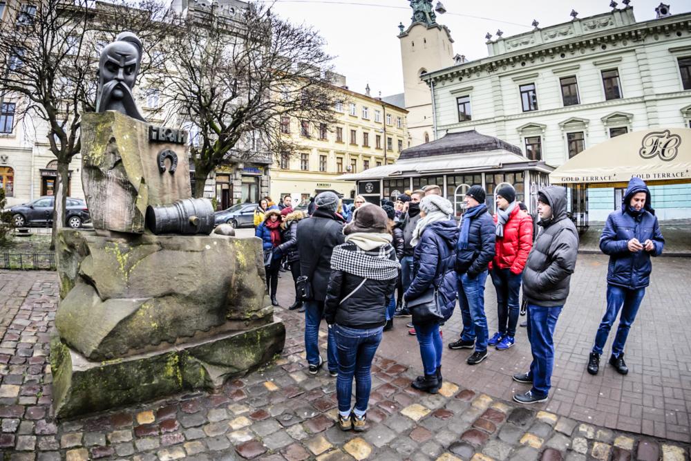 Grupa turystów ogląda pomnik z rzeźbą głowy i elementami symbolizującymi transport w historycznym centrum miasta.