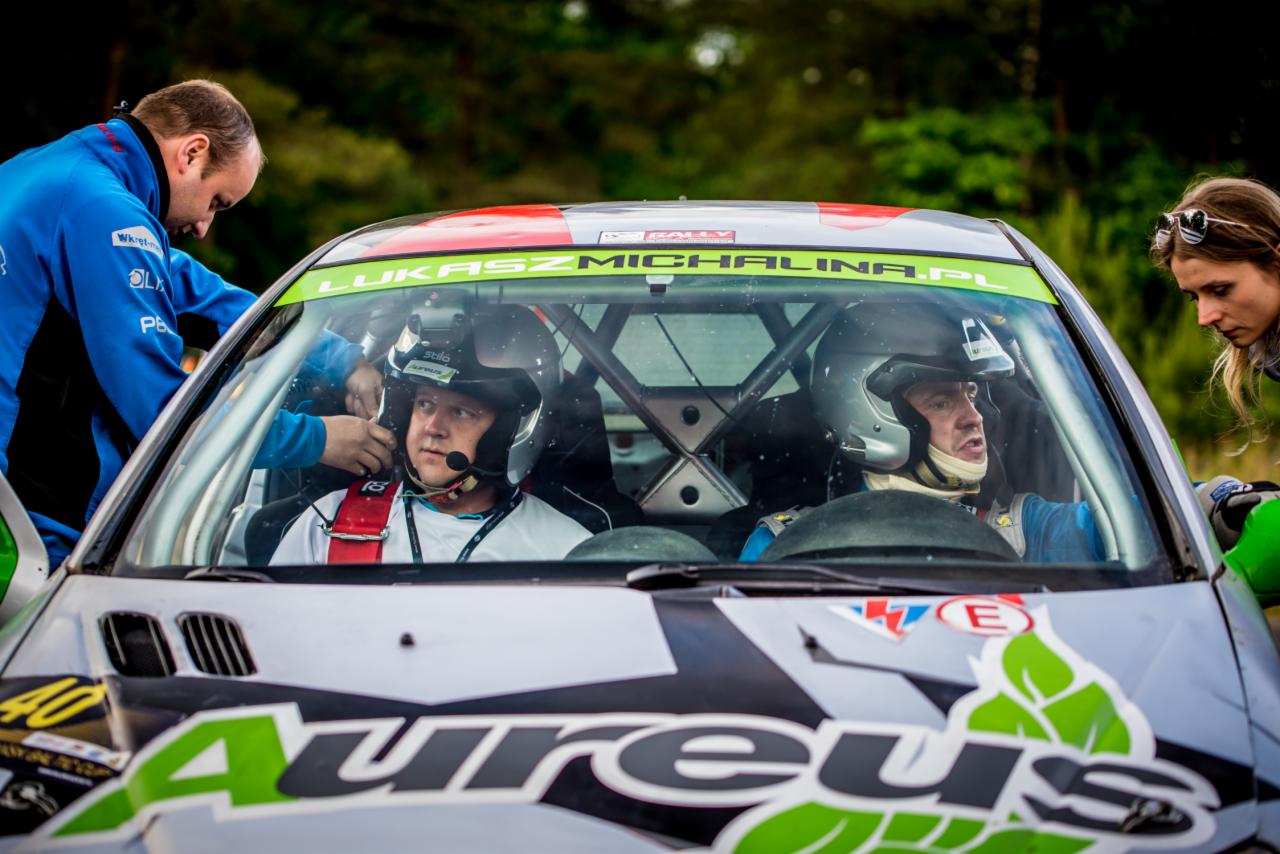 Kierowca rajdowy w kasku, zapinany przez członka zespołu w samochodzie rajdowym z widocznym logotypem sponsora Aureus i numerem startowym 40, przygotowywany do startu w zawodach.