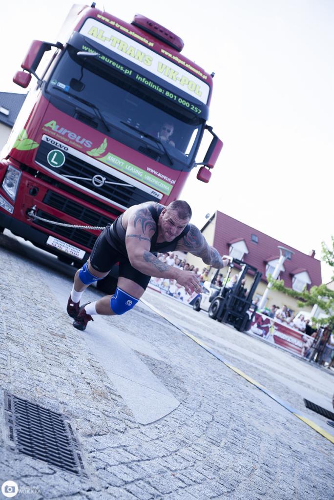 Strongman ciągnie na linie czerwoną ciężarówkę Volvo z logo Aureus na tle publiczności i wózka widłowego.