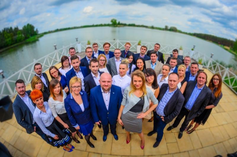 Grupa uśmiechniętych osób w strojach biznesowych, stojąca na tarasie z widokiem na jezioro, sfotografowana z szerokim kątem z góry.