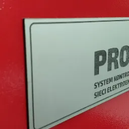 Czerwona obudowa urządzenia z metalową tabliczką informacyjną 'PROTEON: System kontroli zasilania z sieci elektroenergetycznej'.
