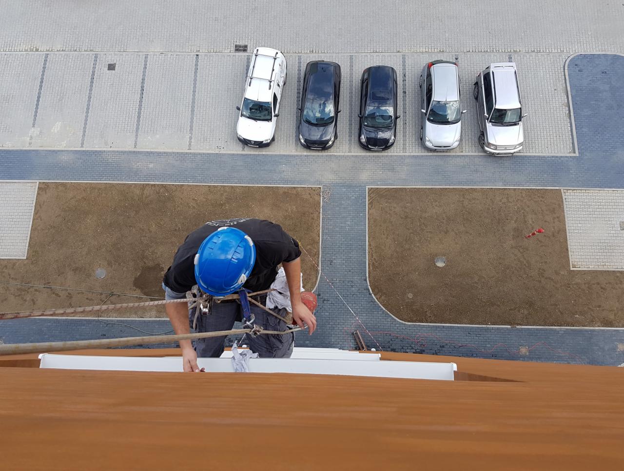 Pracownik w kasku zabezpieczony linami podczas prac na wysokości na brązowej elewacji budynku, widok z góry na parking z samochodami.