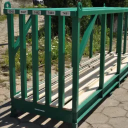 Zielona, metalowa konstrukcja klatkowa z numerowanymi etykietami, stojąca na betonowej kostce brukowej, z drewnianymi belkami wewnątrz, przeznaczona prawdopodobnie do transportu lub przechowywania...