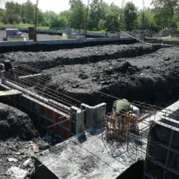 Prace fundamentowe w toku: zbrojenie i szalunki przygotowane pod wylanie betonu, widoczny pracownik przy zbrojeniu, teren budowy w stanie surowym.