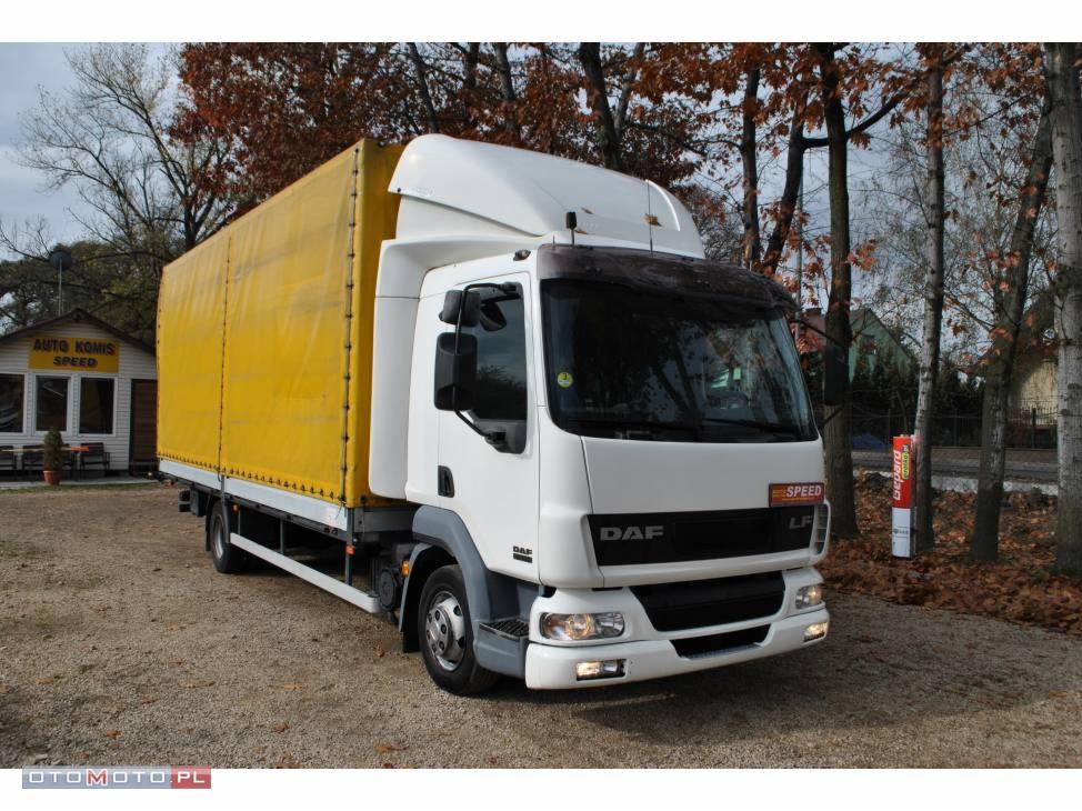 Biały samochód ciężarowy DAF LF z żółtą plandeką, zaparkowany na żwirowym placu przed budynkiem komisu samochodowego w jesiennej scenerii.