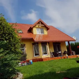 Żółty dom z czerwoną dachówką w trakcie czyszczenia dachu, widoczna różnica między umytą a brudną częścią, na trawniku leżą narzędzia i akcesoria.