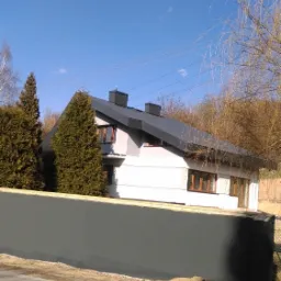 Nowoczesny dom jednorodzinny z grafitowym dachem i jasną elewacją, widoczny zza ciemnoszarego ogrodzenia. Na pierwszym planie asfaltowa droga z cieniami drzew. Błękitne niebo z delikatnymi obłokami...