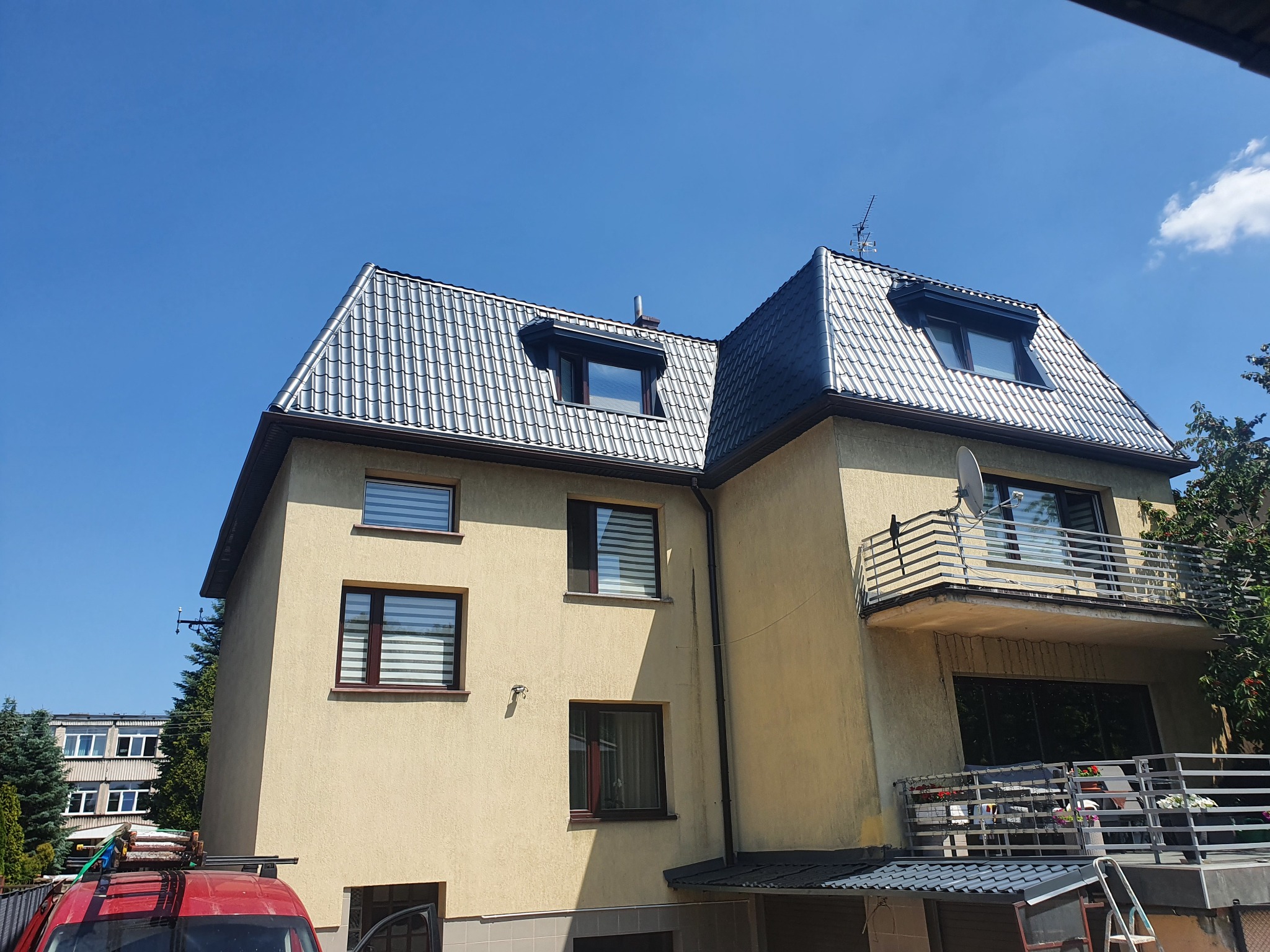 Dwupiętrowy dom z balkonem i nowym, błyszczącym, szarym dachem ceramicznym, widoczny spod kąta, z czerwonym samochodem z bagażnikiem na dachu w lewym dolnym rogu.