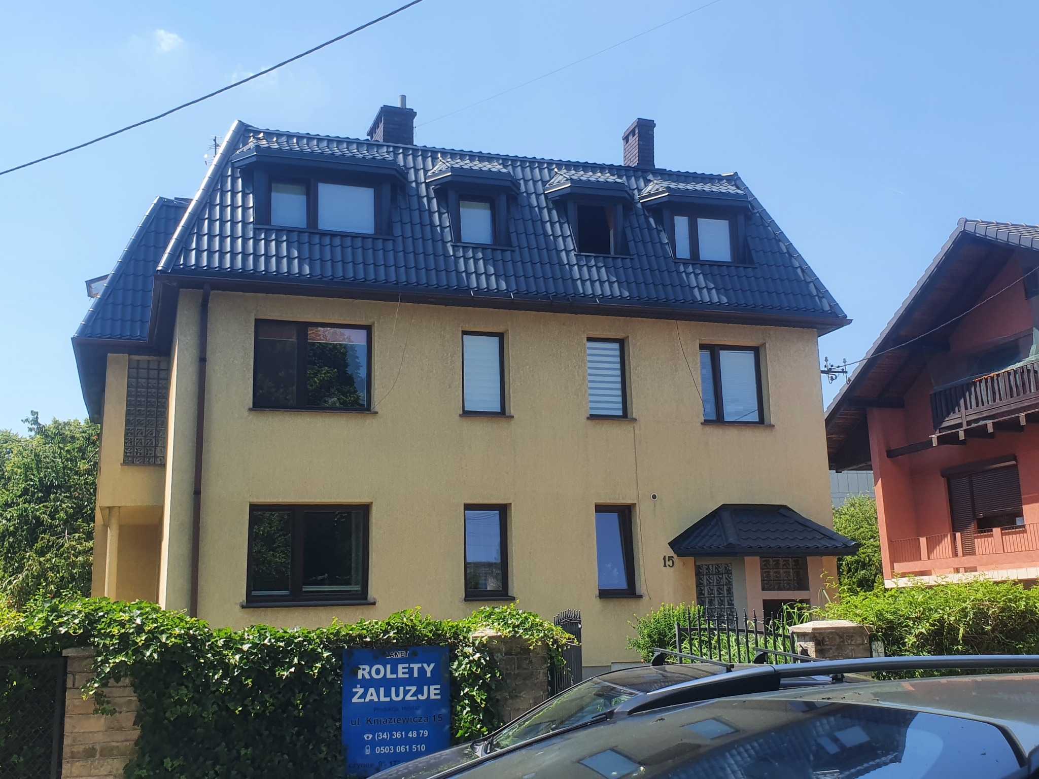 Dwupiętrowy dom z żółtą elewacją i ciemnym, nowym dachem z dachówki bitumicznej, widoczny z poziomu ulicy, z fragmentem samochodu na pierwszym planie i reklamą rolet w tle.