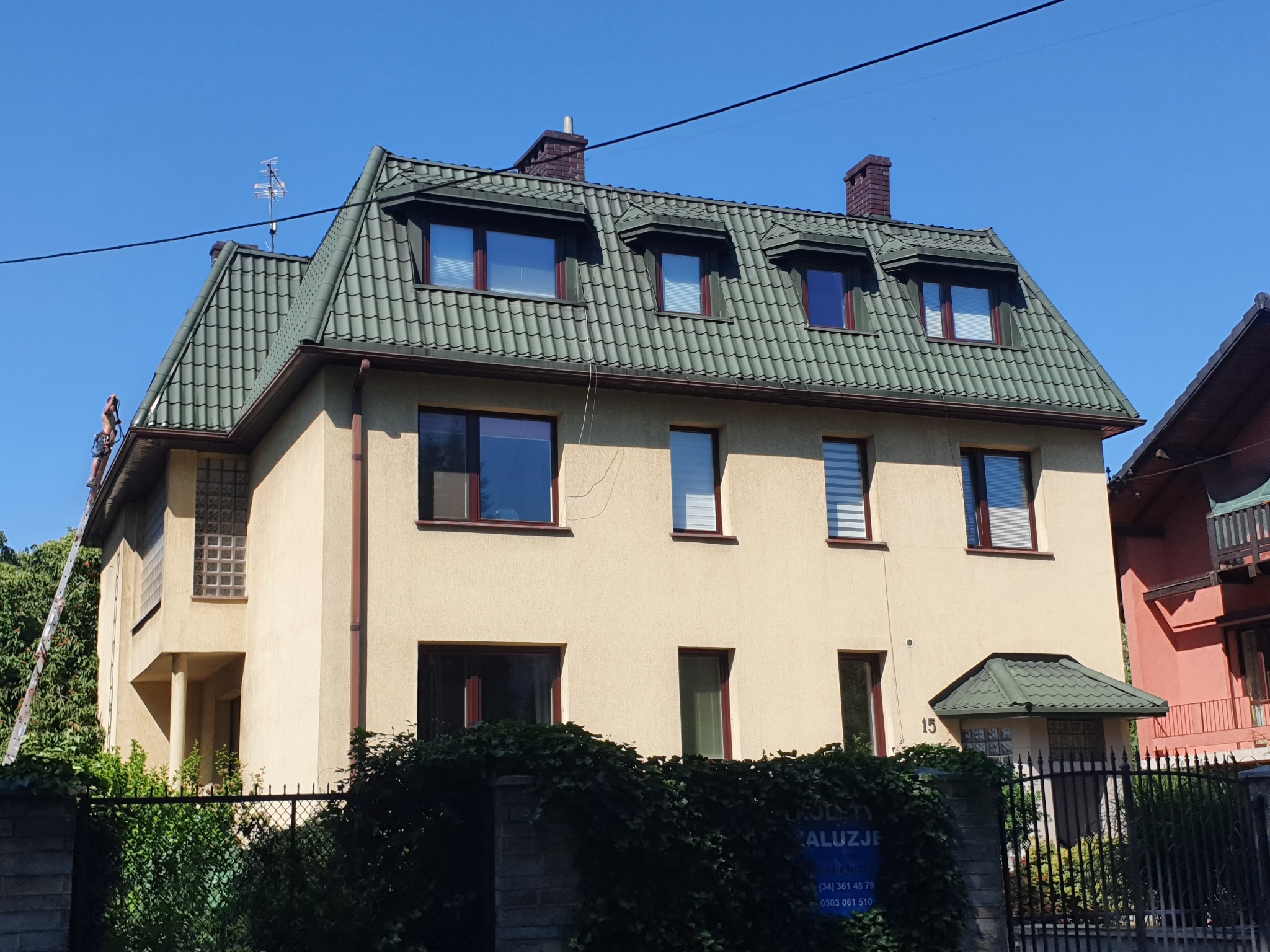 Dwupiętrowy dom z zielonym dachem krytym dachówką, beżową elewacją i pracownikiem na drabinie przy bocznej ścianie.