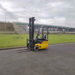 Żółto-czarny wózek widłowy elektryczny XE123 na szarym asfalcie, z widocznym w tle wiaduktem i zielonym trawnikiem.